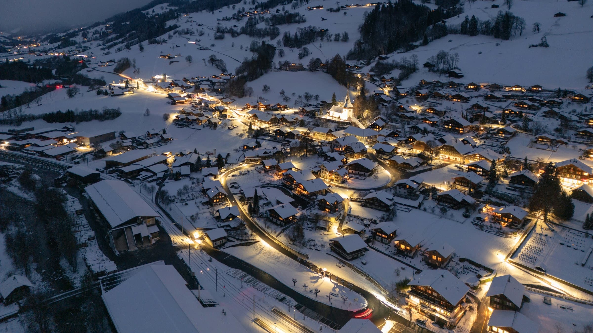 A snow-covered alpine village glowing with warm lights at dusk, chalets and a church spire below the Bernese Oberland peaks.