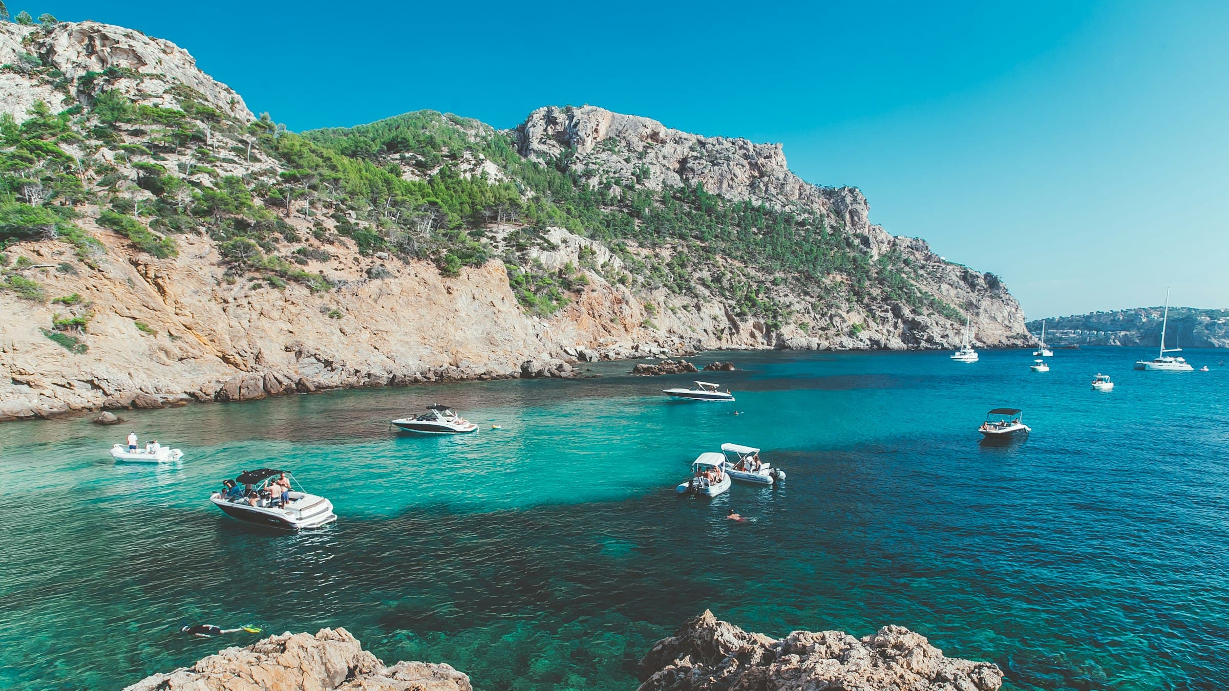 A turquoise Mallorcan cove ringed by pine-topped cliffs, small boats at anchor on the clear water.