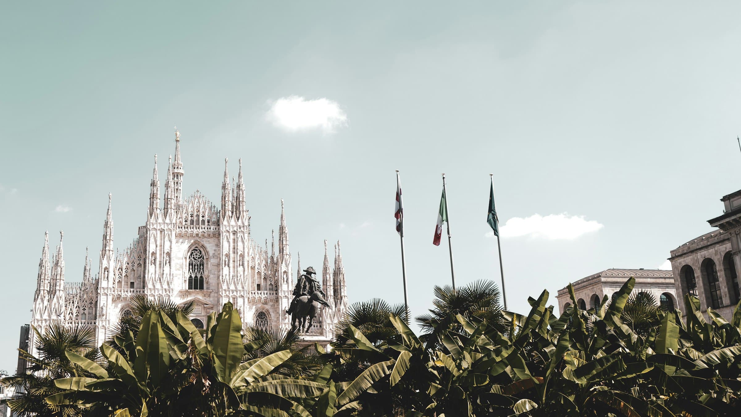 The Gothic spires of Milan's Duomo rising into a pale sky, Italian flags flying alongside.